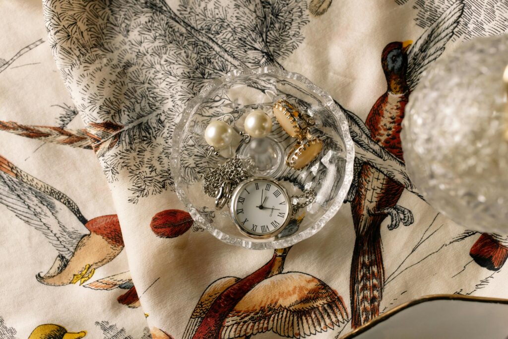 A flat lay of a pocket watch and jewelry in a glass dish over vintage bird-patterned fabric.